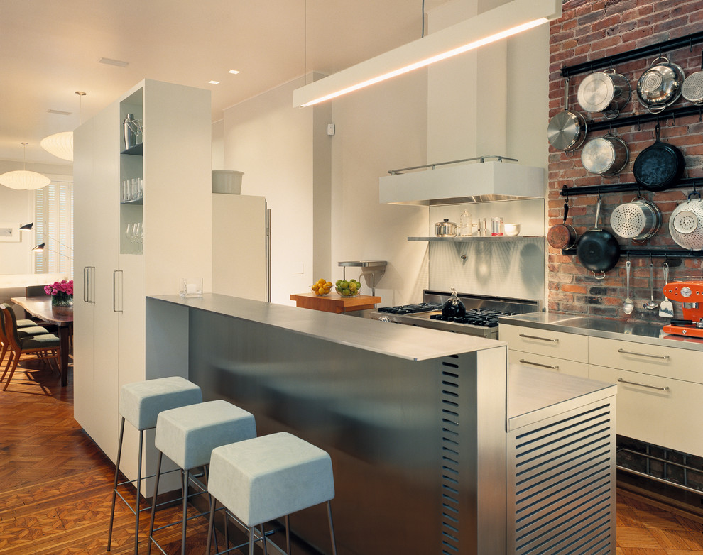 This is an example of a contemporary galley eat-in kitchen in New York with stainless steel appliances, flat-panel cabinets, white cabinets, stainless steel benchtops and glass sheet splashback.