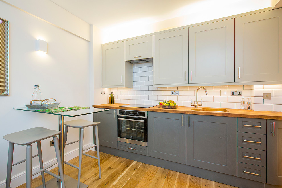 Photo of a mid-sized contemporary single-wall eat-in kitchen in London with an undermount sink, shaker cabinets, grey cabinets, wood benchtops, white splashback, subway tile splashback, stainless steel appliances, light hardwood floors and no island.