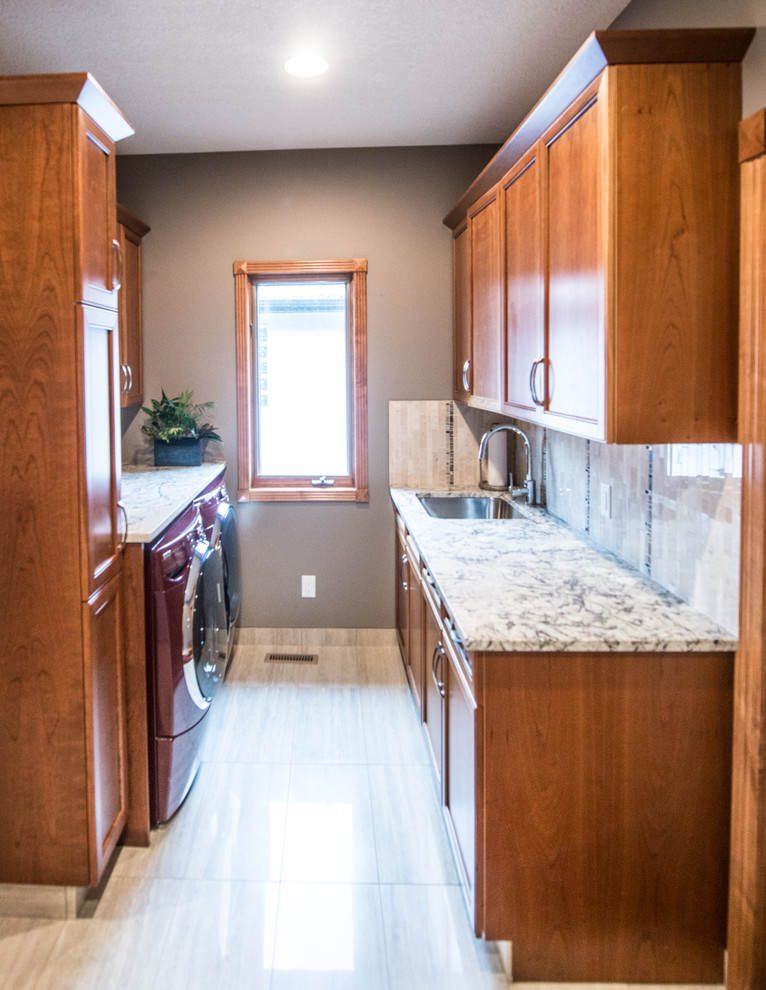 Design ideas for a mid-sized transitional galley dedicated laundry room in Calgary with an undermount sink, shaker cabinets, medium wood cabinets, granite benchtops, grey walls, travertine floors and a side-by-side washer and dryer.