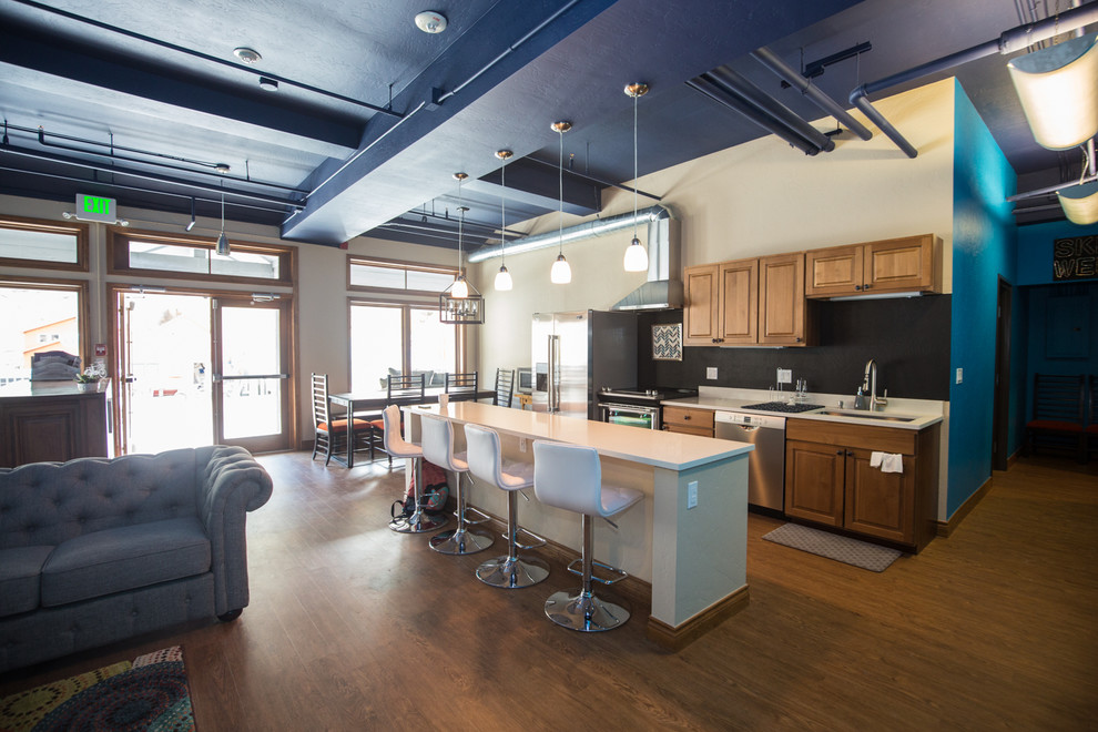 Photo of a mid-sized industrial galley open plan kitchen in Denver with an undermount sink, raised-panel cabinets, dark wood cabinets, solid surface benchtops, black splashback, stone slab splashback, stainless steel appliances, dark hardwood floors, with island and brown floor.