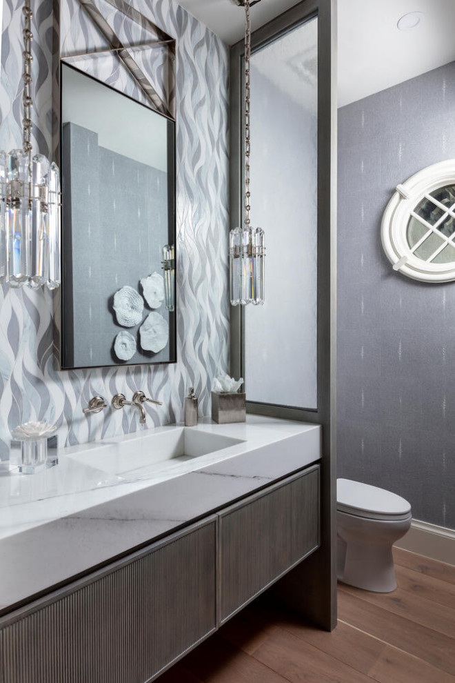 Expansive transitional powder room in Miami with furniture-like cabinets, brown cabinets, a one-piece toilet, gray tile, glass tile, grey walls, medium hardwood floors, an integrated sink, marble benchtops, brown floor, white benchtops, a floating vanity and wallpaper.