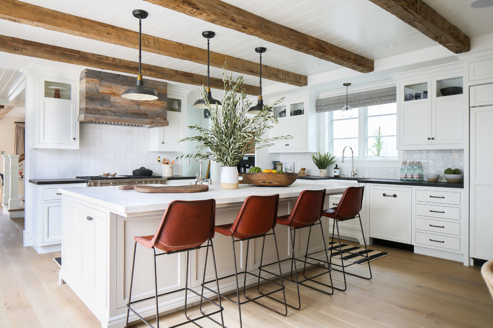 This is an example of a beach style l-shaped open plan kitchen in Los Angeles with a farmhouse sink, recessed-panel cabinets, white cabinets, white splashback, panelled appliances, medium hardwood floors, with island and brown floor.