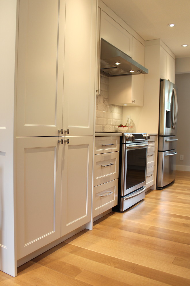 Example of a large transitional l-shaped light wood floor eat-in kitchen design with an undermount sink, shaker cabinets, white cabinets, quartz countertops, white backsplash, ceramic backsplash, stainless steel appliances and an island