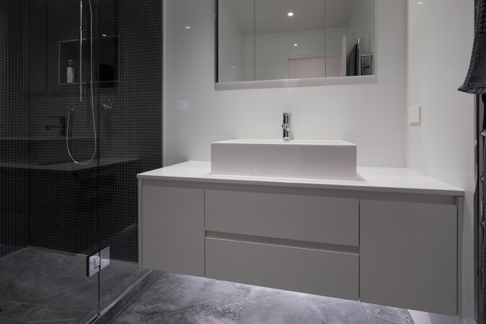 Small contemporary bathroom in Auckland with a trough sink, flat-panel cabinets, white cabinets, solid surface benchtops, a corner shower, a one-piece toilet, white tile, mosaic tile, white walls and ceramic floors.
