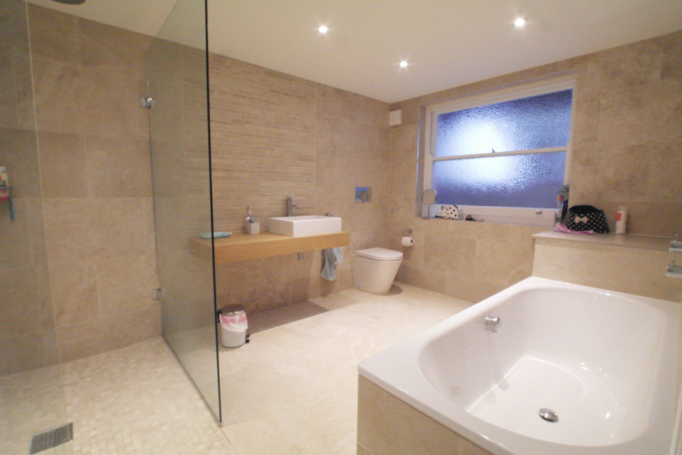 Photo of a mid-sized contemporary master bathroom in London with flat-panel cabinets, grey cabinets, a drop-in tub, an open shower, a one-piece toilet, beige tile, porcelain tile, porcelain floors, a wall-mount sink and tile benchtops.