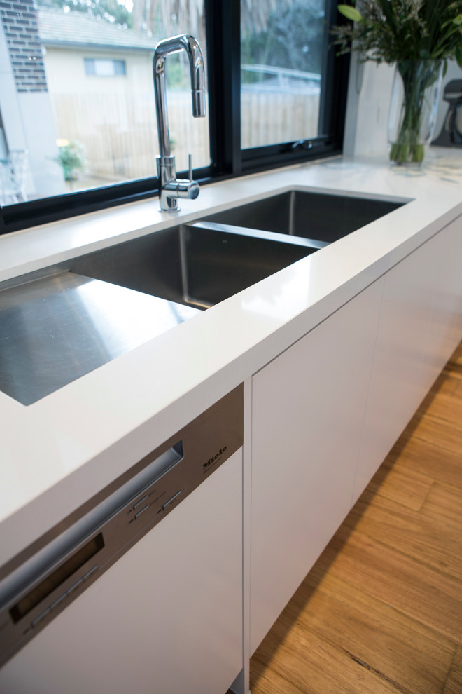 Large contemporary l-shaped eat-in kitchen in Melbourne with a double-bowl sink, flat-panel cabinets, white cabinets, quartz benchtops, window splashback, white appliances, medium hardwood floors and with island.