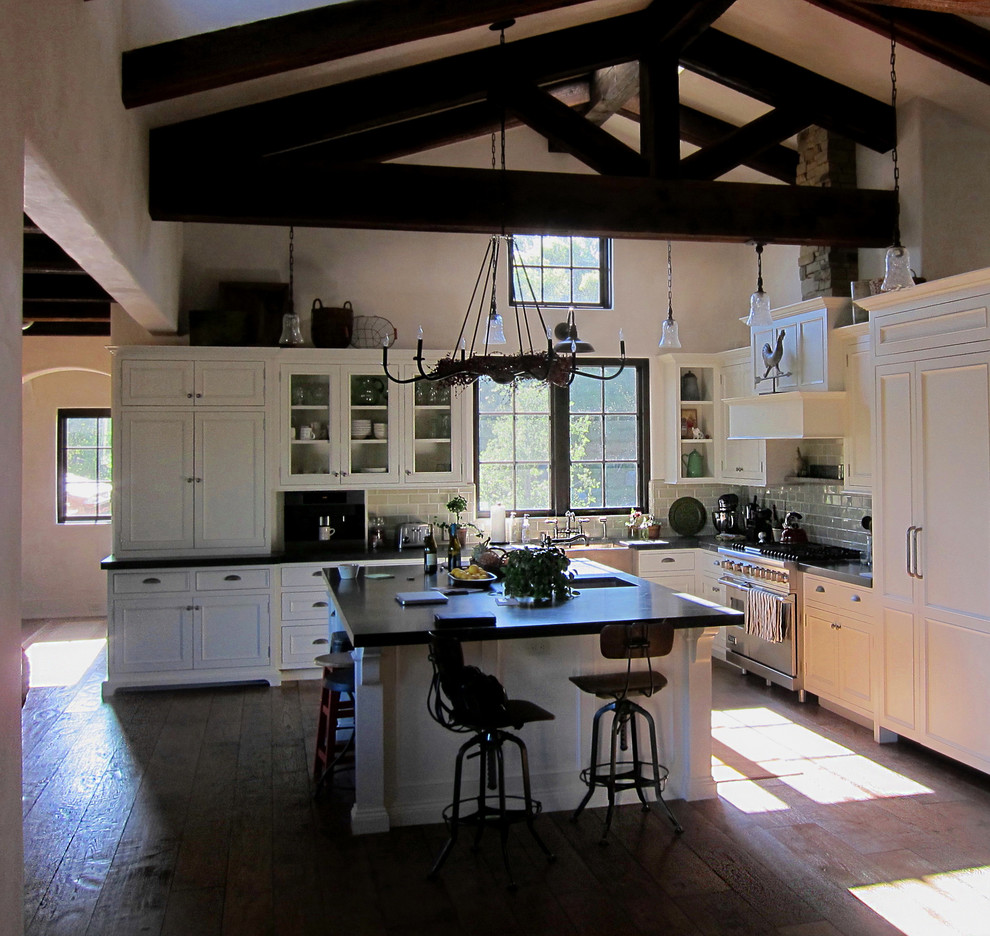 Design ideas for a large country l-shaped open plan kitchen in Santa Barbara with a farmhouse sink, recessed-panel cabinets, white cabinets, soapstone benchtops, green splashback, stainless steel appliances, medium hardwood floors, with island, subway tile splashback, brown floor and black benchtop.