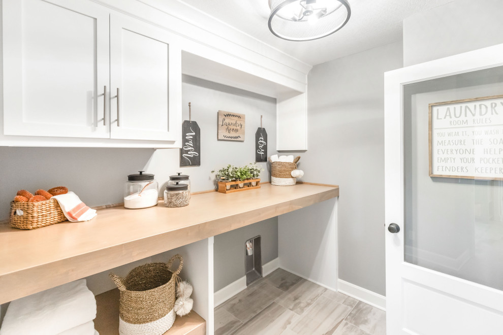 Photo of a large beach style single-wall dedicated laundry room in Kansas City with shaker cabinets, white cabinets, wood benchtops, grey walls, porcelain floors, a side-by-side washer and dryer, grey floor and brown benchtop.