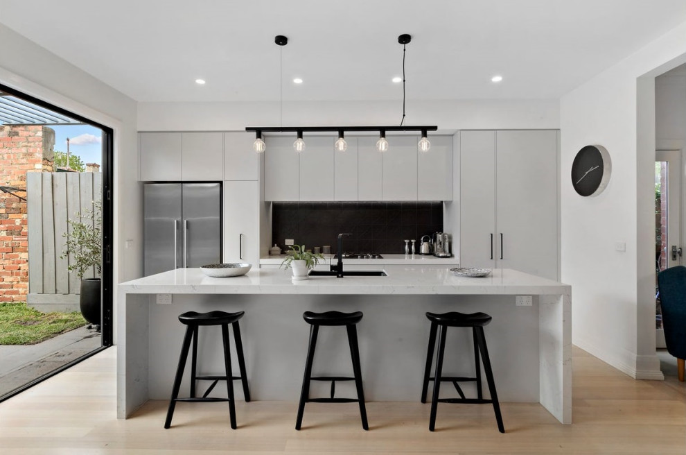Photo of a contemporary galley kitchen/diner in Melbourne with a submerged sink, flat-panel cabinets, white cabinets, engineered stone countertops, black splashback, metal splashback, black appliances, light hardwood flooring, an island, grey floors and white worktops.