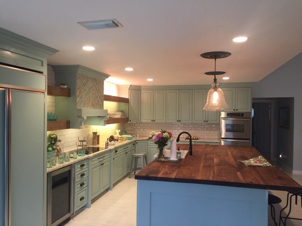 This is an example of a large country l-shaped eat-in kitchen in Miami with a farmhouse sink, raised-panel cabinets, green cabinets, wood benchtops, white splashback, subway tile splashback, panelled appliances, with island, white floor and brown benchtop.
