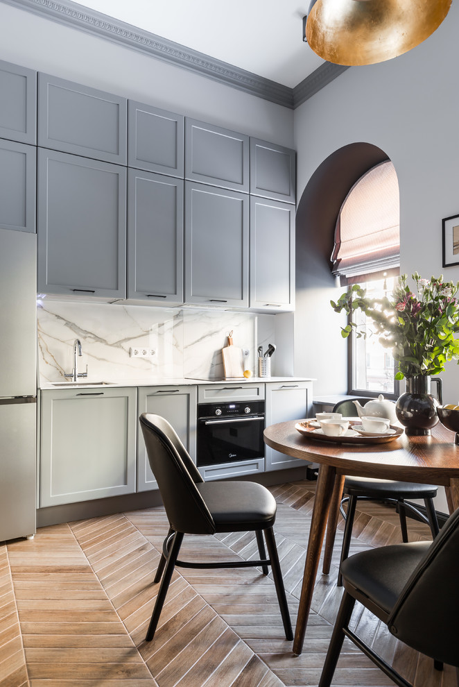 This is an example of a transitional single-wall eat-in kitchen in Saint Petersburg with an undermount sink, grey cabinets, solid surface benchtops, white splashback, ceramic splashback, black appliances, ceramic floors, beige floor, white benchtop and recessed-panel cabinets.