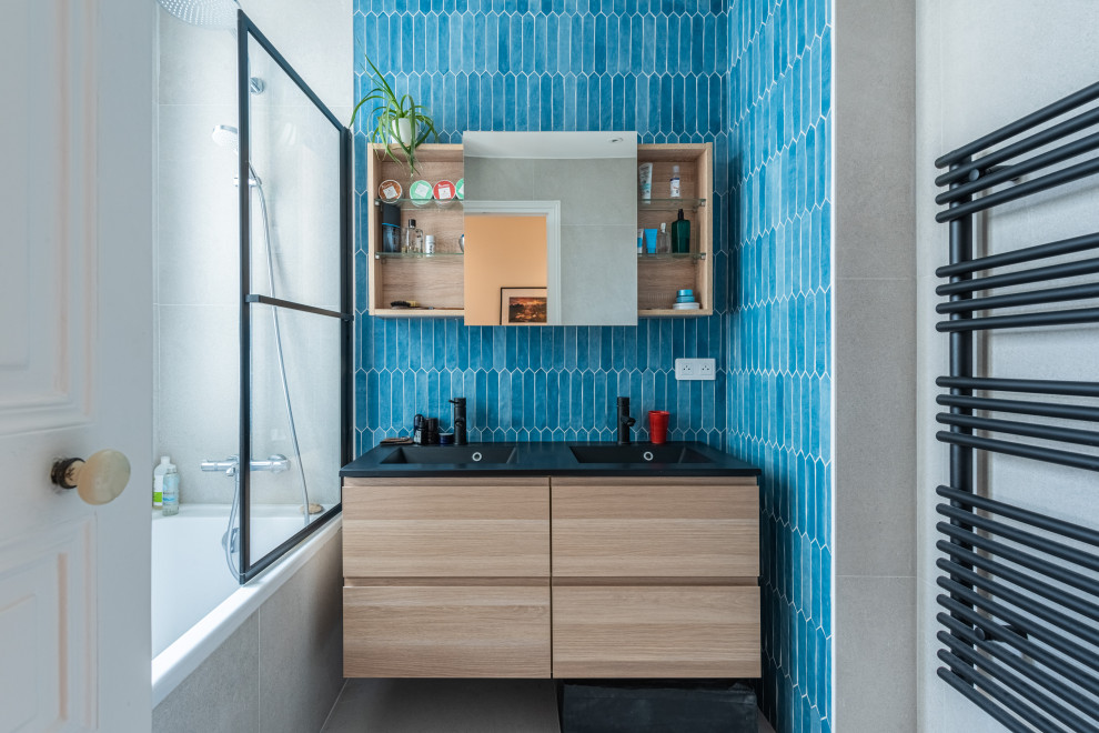 Medium sized contemporary ensuite bathroom in Paris with flat-panel cabinets, light wood cabinets, a submerged bath, a shower/bath combination, a two-piece toilet, blue tiles, ceramic tiles, blue walls, ceramic flooring, a console sink, a hinged door, black worktops, double sinks and a floating vanity unit.