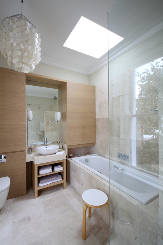 Photo of a country master bathroom in Dublin with flat-panel cabinets, beige cabinets, a drop-in tub, a shower/bathtub combo, a wall-mount toilet, beige tile, limestone, beige walls, limestone floors, a vessel sink, limestone benchtops, beige floor, a hinged shower door, beige benchtops, a single vanity and a built-in vanity.