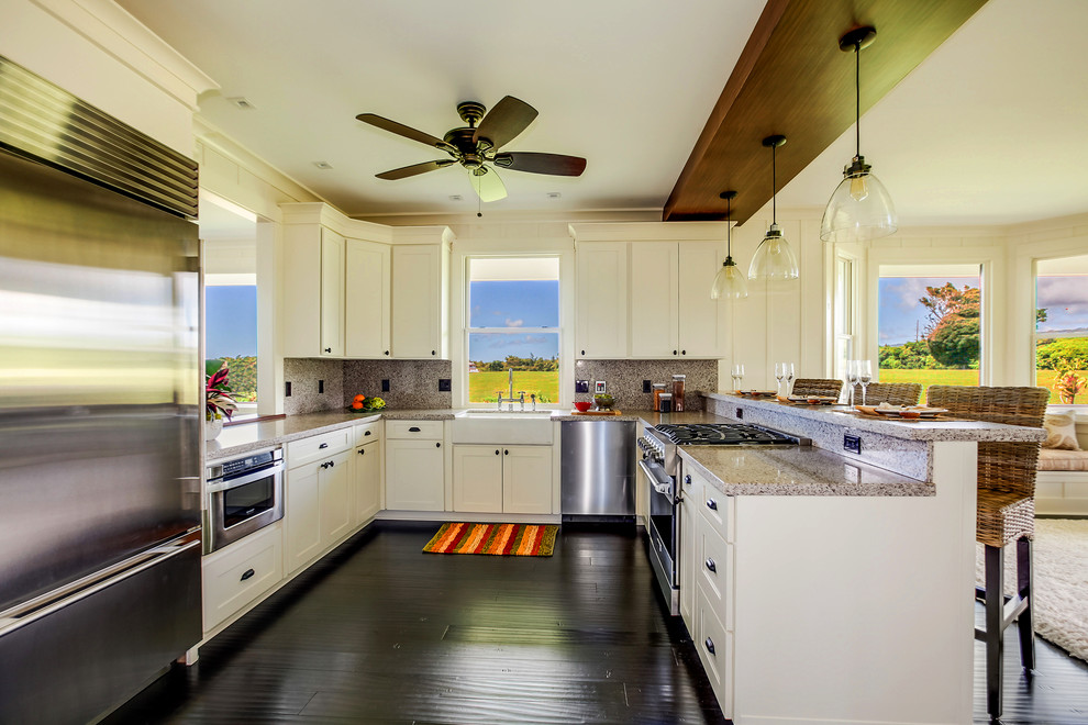 Photo of a mid-sized beach style u-shaped open plan kitchen in Hawaii with a farmhouse sink, shaker cabinets, white cabinets, granite benchtops, grey splashback, stone slab splashback, stainless steel appliances, bamboo floors, a peninsula and black floor.