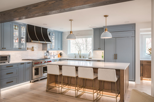 Modern kitchen with blue-gray shaker kitchen cabinet run, brass hardware, glass-front uppers, panel-ready fridge, and a walnut island with white quartz and upholstered stools under brass dome pendants.
