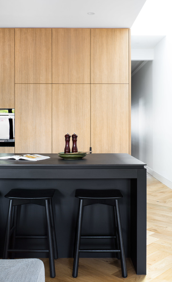 Photo of a mid-sized contemporary galley eat-in kitchen in Melbourne with a drop-in sink, flat-panel cabinets, medium wood cabinets, concrete benchtops, black appliances, light hardwood floors, with island, brown floor and black benchtop.