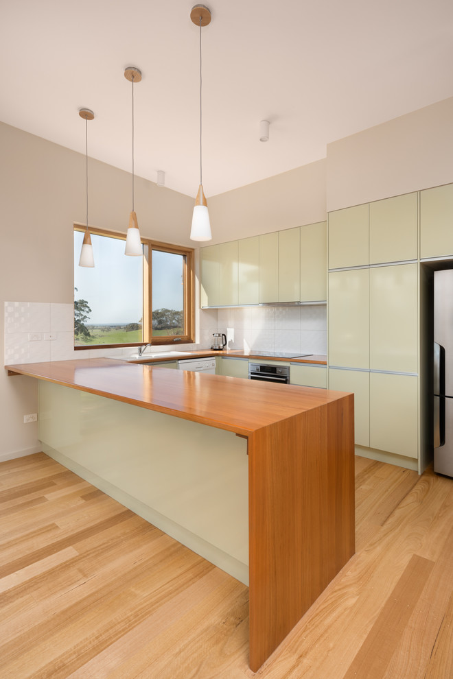 Design ideas for a mid-sized contemporary l-shaped eat-in kitchen in Other with green cabinets, wood benchtops, white splashback, ceramic splashback, light hardwood floors, a drop-in sink, flat-panel cabinets, stainless steel appliances, a peninsula, beige floor and brown benchtop.