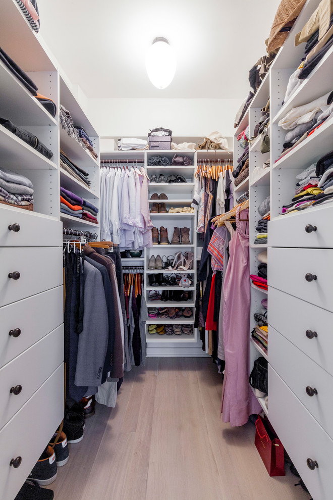 This is an example of a mid-sized transitional gender-neutral walk-in wardrobe in New York with flat-panel cabinets, white cabinets, light hardwood floors and beige floor.