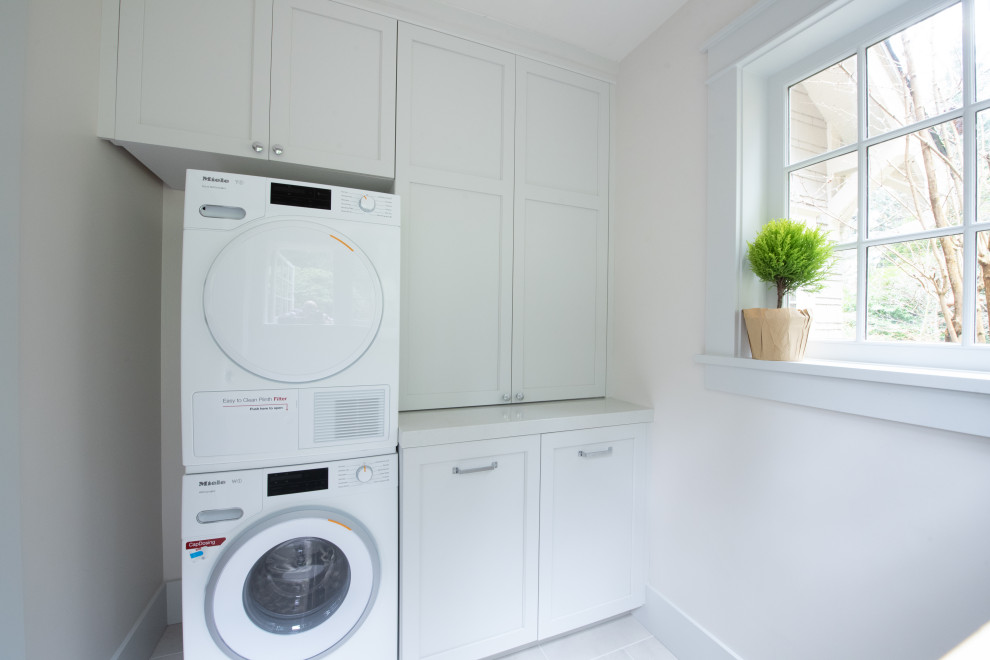 This is an example of a small traditional galley dedicated laundry room in Seattle with recessed-panel cabinets, white cabinets, quartz benchtops, white walls, porcelain floors, a stacked washer and dryer, white floor and white benchtop.