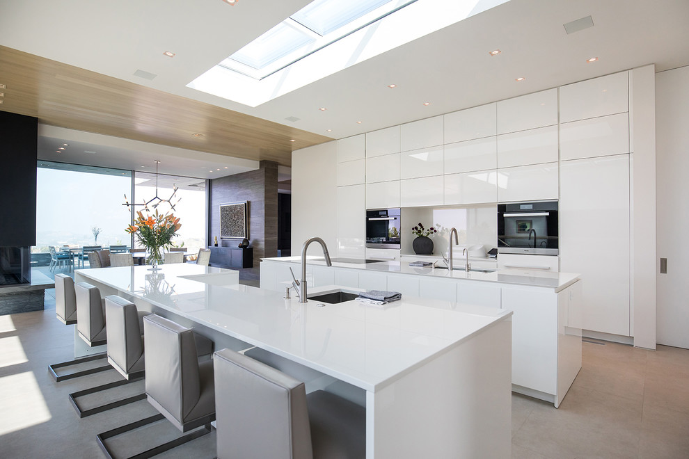 This is an example of an expansive contemporary galley eat-in kitchen in Los Angeles with an undermount sink, flat-panel cabinets, white cabinets, white splashback, multiple islands, white benchtop, black appliances, porcelain floors and white floor.