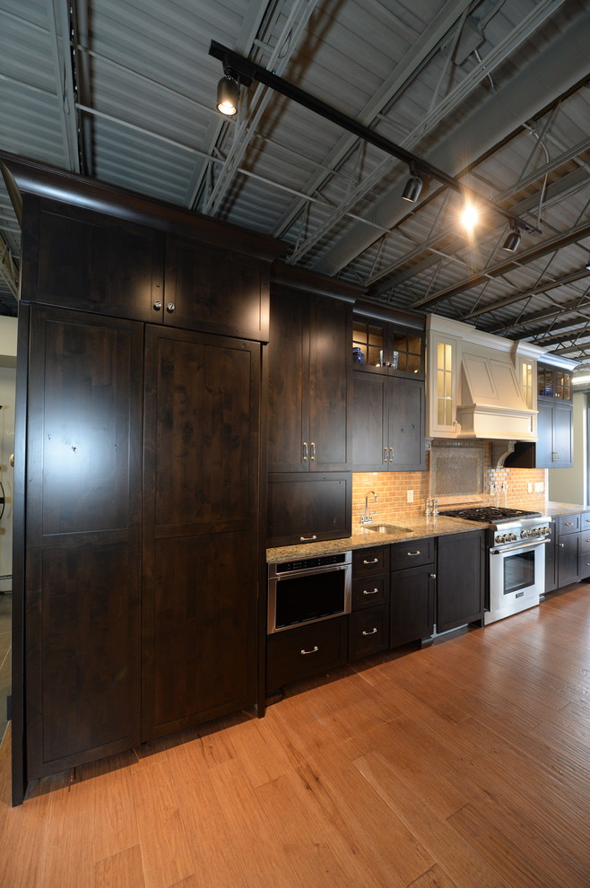 Photo of an industrial kitchen in Cleveland with an undermount sink, shaker cabinets, beige splashback, stone tile splashback, stainless steel appliances, porcelain floors, dark wood cabinets and granite benchtops.