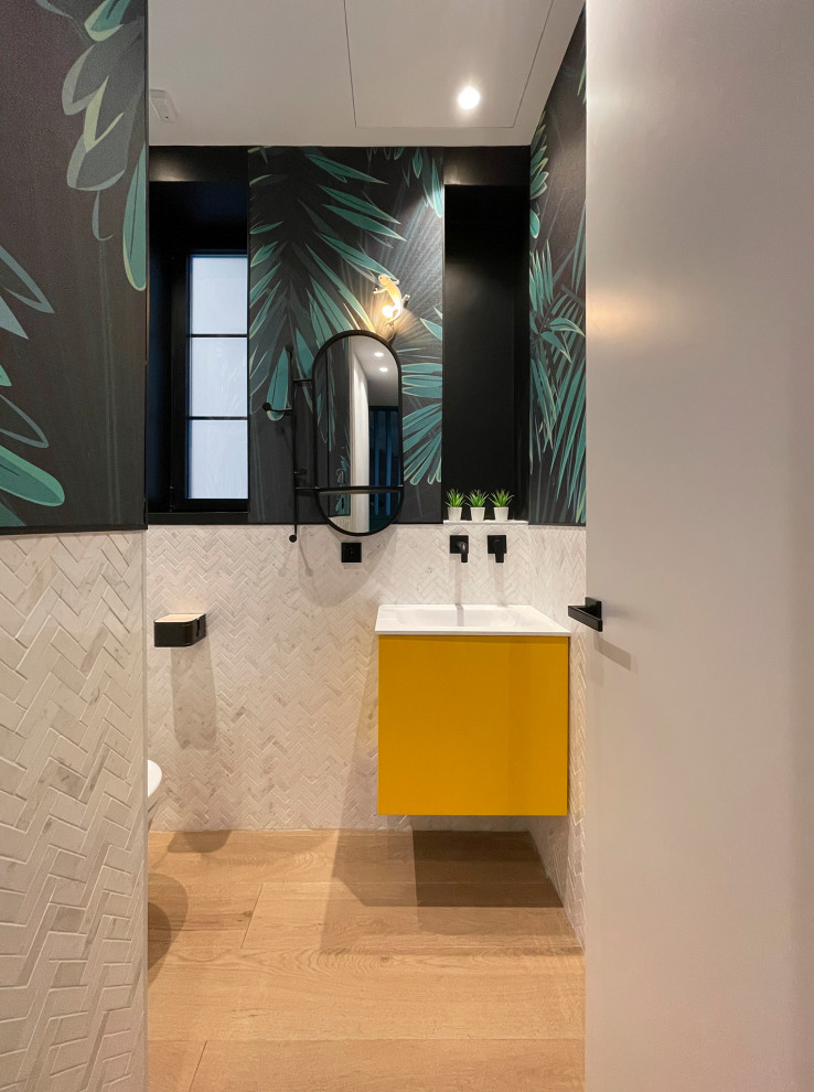 This is an example of a small tropical bathroom in Other with flat-panel cabinets, yellow cabinets, a wall-mount toilet, white tile, marble, white walls, medium hardwood floors, a drop-in sink, solid surface benchtops, brown floor, white benchtops, a single vanity, a built-in vanity and wallpaper.