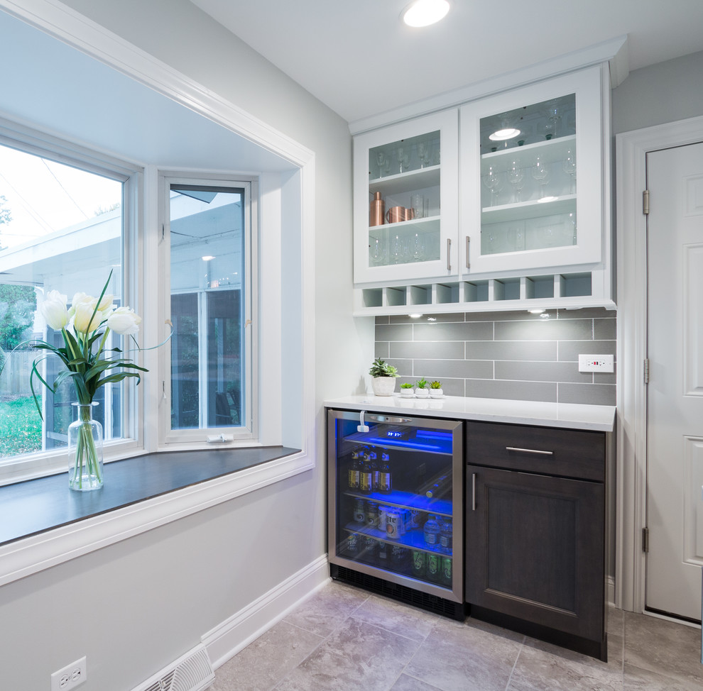 This is an example of a small transitional single-wall wet bar in Chicago with white cabinets, quartz benchtops, grey splashback, porcelain splashback, porcelain floors, glass-front cabinets, beige floor and white benchtop.