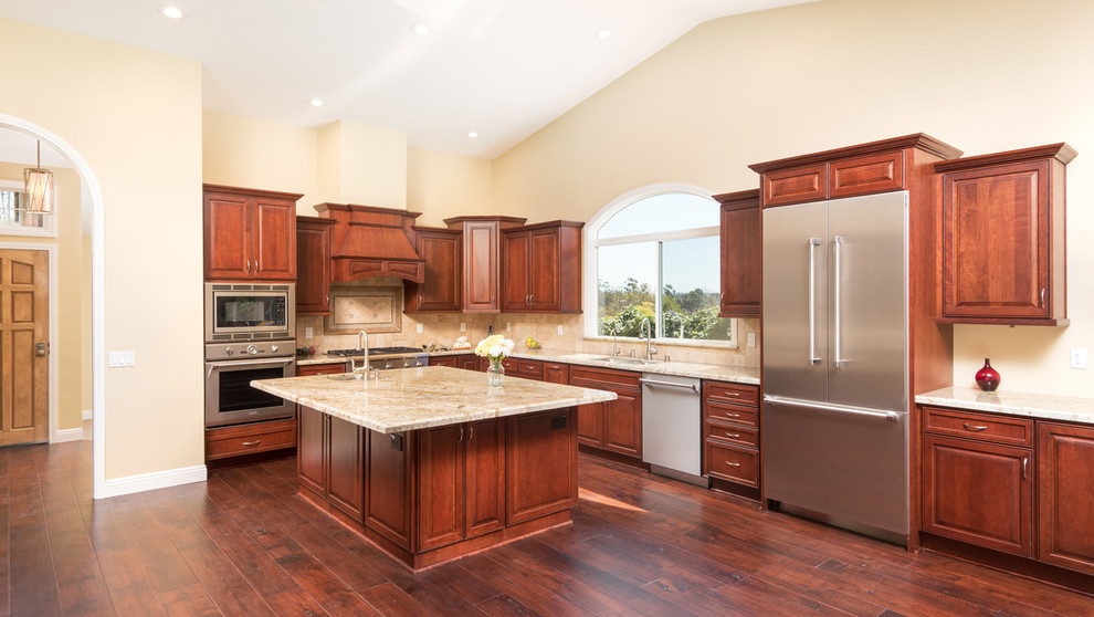 This is an example of an expansive traditional l-shaped open plan kitchen in San Diego with raised-panel cabinets, dark wood cabinets, granite benchtops, beige splashback, stone tile splashback, stainless steel appliances, dark hardwood floors, with island, an undermount sink, beige floor, beige benchtop and vaulted.