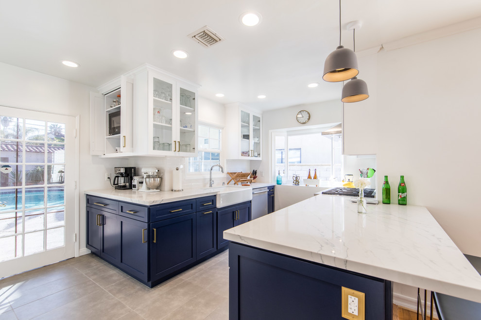 This is an example of a mid-sized transitional u-shaped open plan kitchen in Los Angeles with a farmhouse sink, shaker cabinets, blue cabinets, marble benchtops, stainless steel appliances, porcelain floors, a peninsula, beige floor and beige benchtop.
