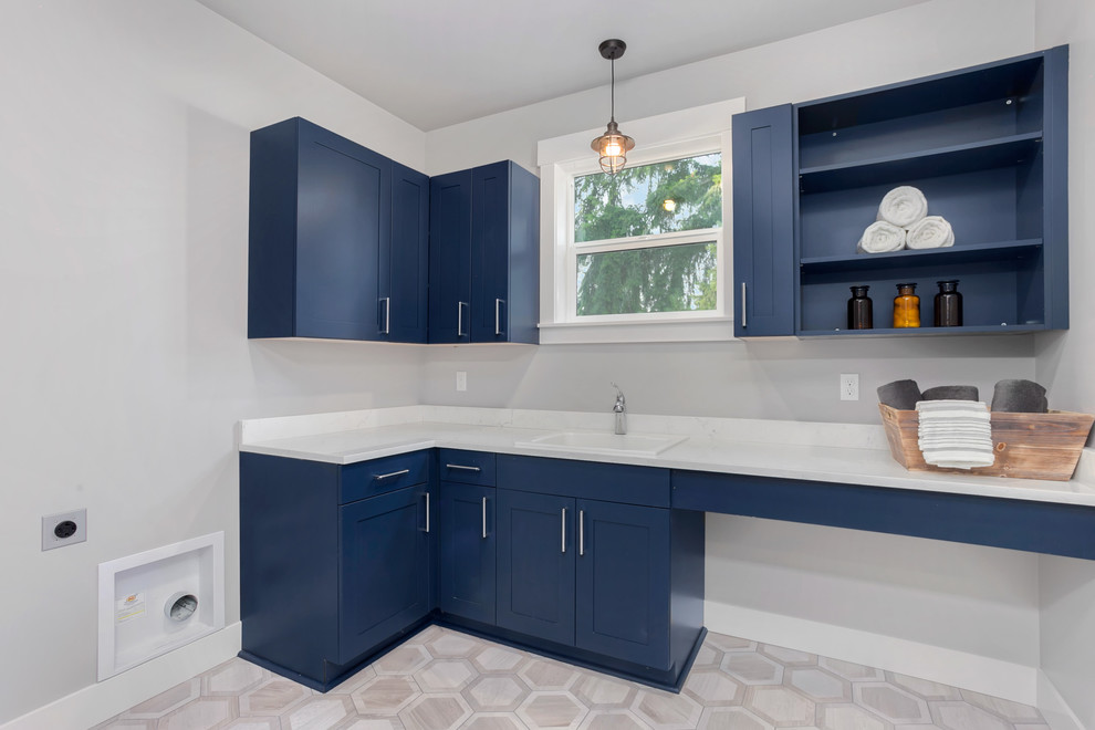 This is an example of a mid-sized transitional l-shaped dedicated laundry room in Seattle with a drop-in sink, shaker cabinets, blue cabinets, quartz benchtops, grey walls, ceramic floors, a side-by-side washer and dryer and grey floor.