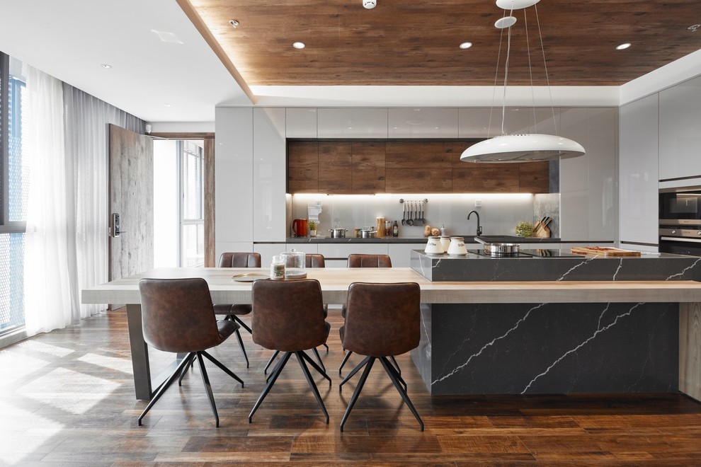 Photo of a contemporary u-shaped eat-in kitchen in Hawaii with flat-panel cabinets, medium wood cabinets, grey splashback, glass sheet splashback, stainless steel appliances, medium hardwood floors, brown floor, grey benchtop and with island.