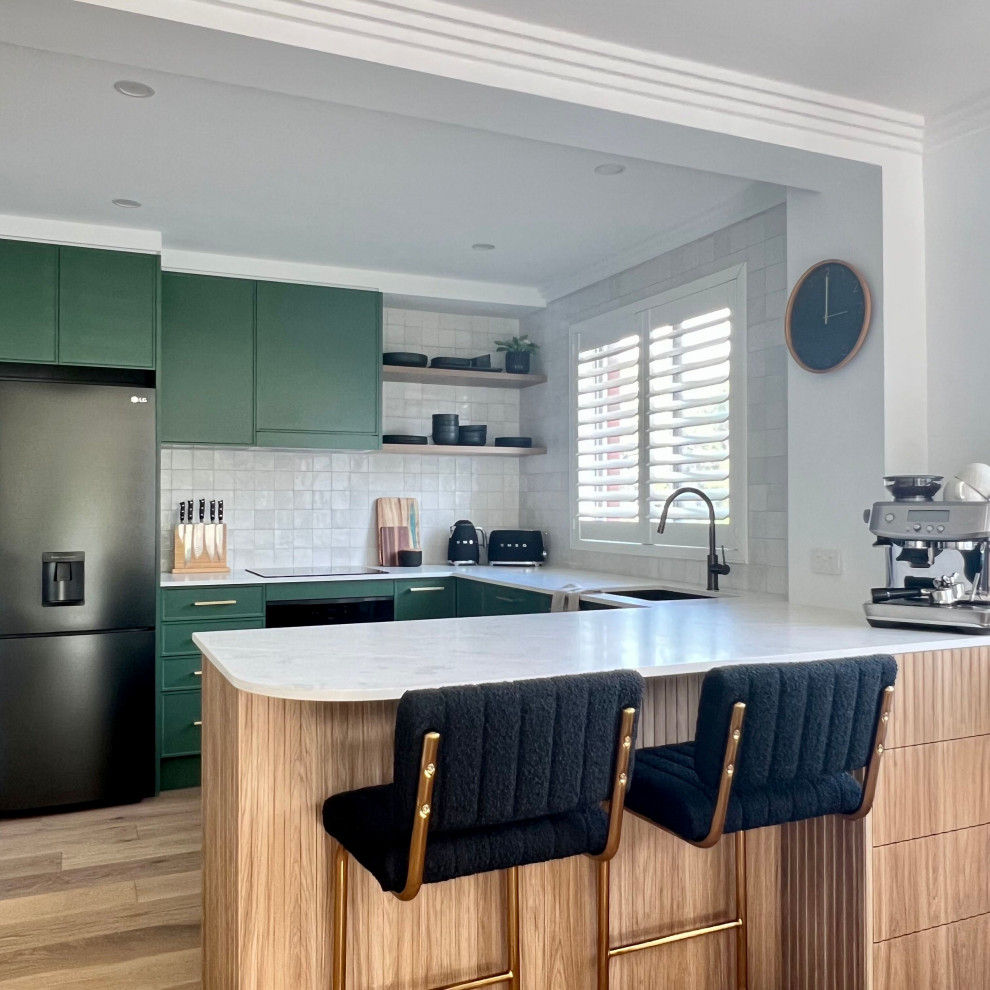 This is an example of a mid-sized contemporary u-shaped open plan kitchen in Sydney with an undermount sink, recessed-panel cabinets, green cabinets, quartz benchtops, yellow splashback, porcelain splashback, black appliances, medium hardwood floors, a peninsula, brown floor and white benchtop.