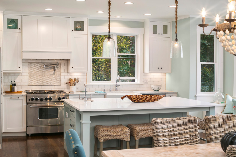 Mid-sized beach style l-shaped eat-in kitchen in Seattle with a farmhouse sink, shaker cabinets, white cabinets, marble benchtops, white splashback, porcelain splashback, stainless steel appliances, dark hardwood floors and with island.