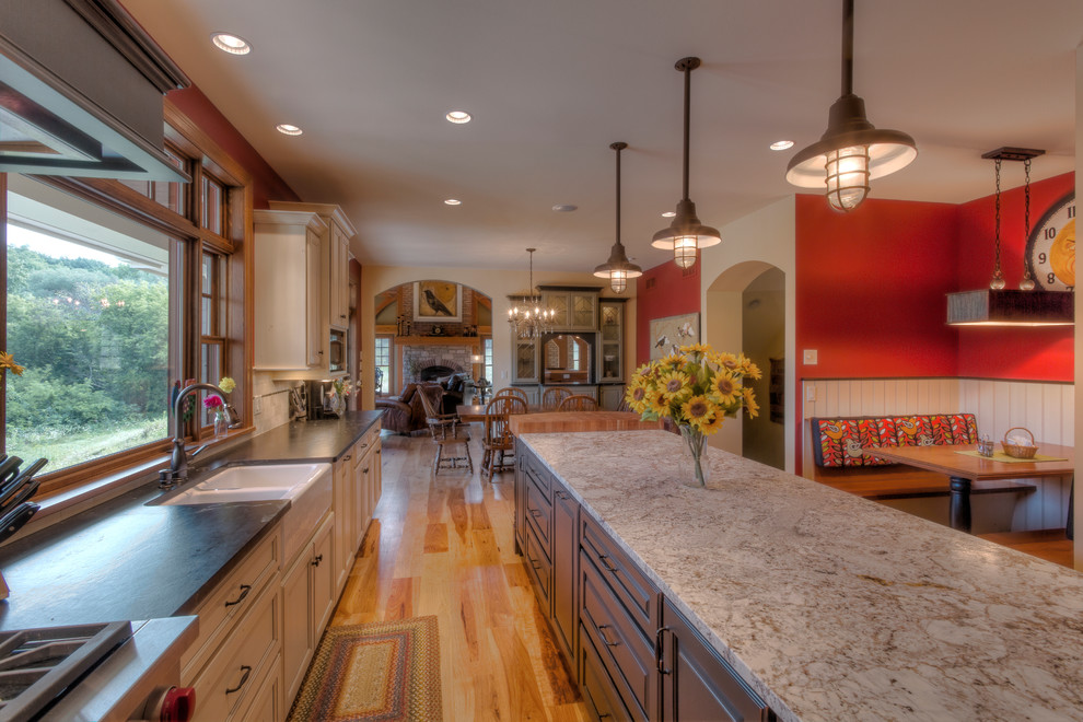 Example of a mountain style l-shaped medium tone wood floor eat-in kitchen design with a farmhouse sink, raised-panel cabinets, white cabinets, granite countertops, white backsplash, ceramic backsplash, stainless steel appliances and an island