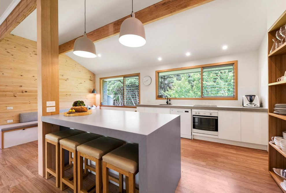 Photo of a beach style kitchen in Melbourne with a single-bowl sink, flat-panel cabinets, white cabinets, white appliances, medium hardwood floors, with island, grey benchtop and exposed beam.