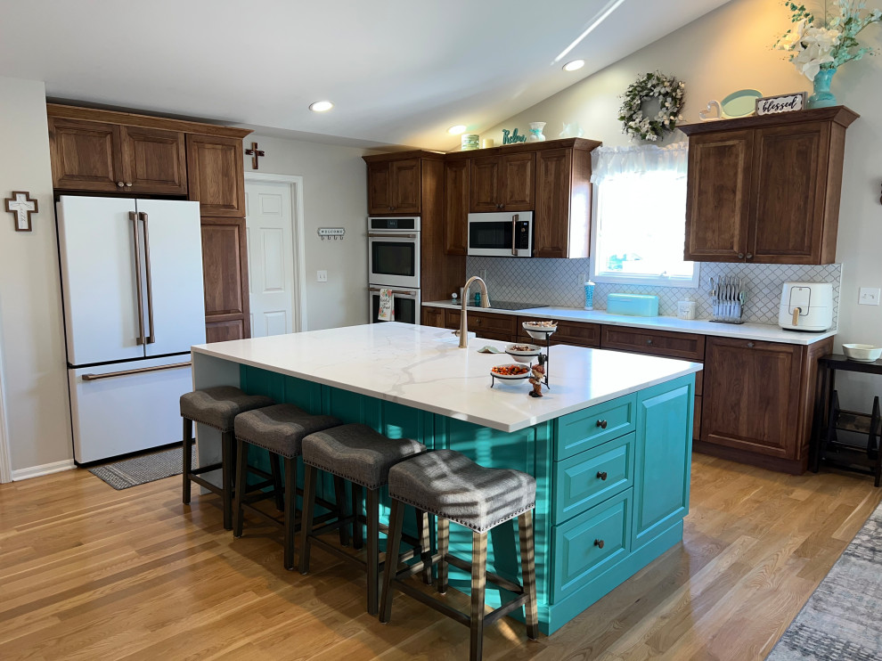 This is an example of a mid-sized transitional u-shaped eat-in kitchen in Detroit with an undermount sink, raised-panel cabinets, brown cabinets, quartz benchtops, white splashback, ceramic splashback, medium hardwood floors, with island, brown floor, white benchtop, vaulted and white appliances.