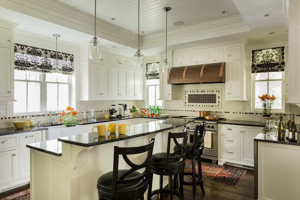Photo of a traditional u-shaped eat-in kitchen in Boston with white cabinets, white splashback, subway tile splashback, stainless steel appliances, with island, brown floor, black benchtop, a farmhouse sink, recessed-panel cabinets and dark hardwood floors.