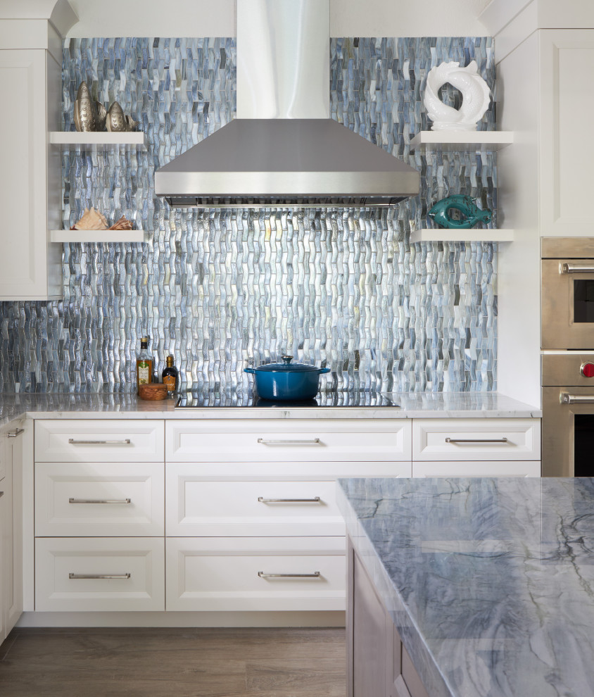 Photo of a large beach style l-shaped kitchen in Other with a farmhouse sink, recessed-panel cabinets, white cabinets, quartzite benchtops, multi-coloured splashback, glass tile splashback, stainless steel appliances, porcelain floors, with island, grey floor and blue benchtop.