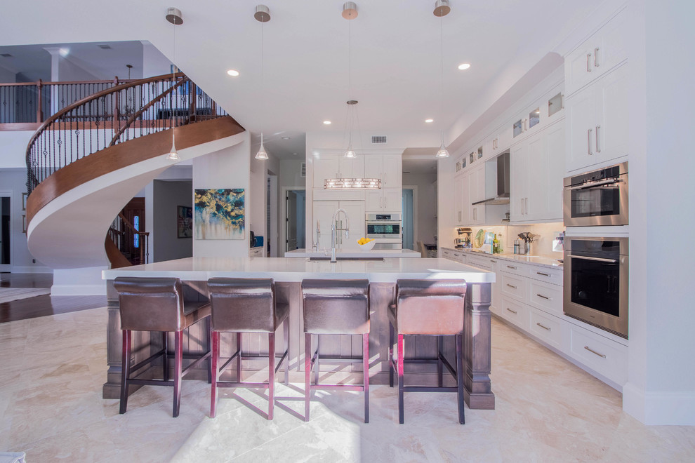 Example of a huge transitional l-shaped porcelain tile and beige floor open concept kitchen design with a farmhouse sink, shaker cabinets, white cabinets, quartz countertops, white backsplash, porcelain backsplash, stainless steel appliances and two islands