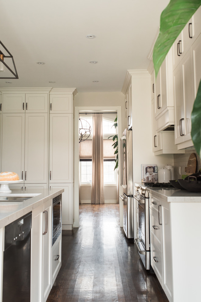 Example of a transitional medium tone wood floor and brown floor kitchen design in Toronto with an undermount sink, recessed-panel cabinets, white cabinets, quartz countertops, white backsplash, porcelain backsplash, stainless steel appliances and an island