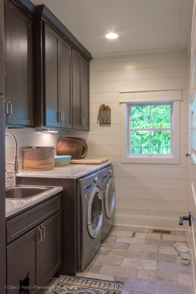 This is an example of a mid-sized country single-wall dedicated laundry room in Minneapolis with a drop-in sink, flat-panel cabinets, distressed cabinets, solid surface benchtops, white walls, ceramic floors, a side-by-side washer and dryer, grey floor and grey benchtop.