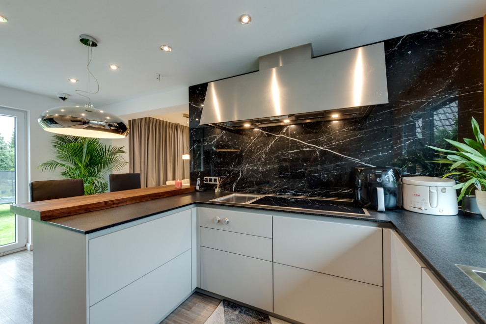 Large traditional grey and white u-shaped open plan kitchen in Hamburg with an integrated sink, flat-panel cabinets, white cabinets, composite countertops, black splashback, marble splashback, black appliances, terrazzo flooring, a breakfast bar, multi-coloured floors, black worktops and a drop ceiling.