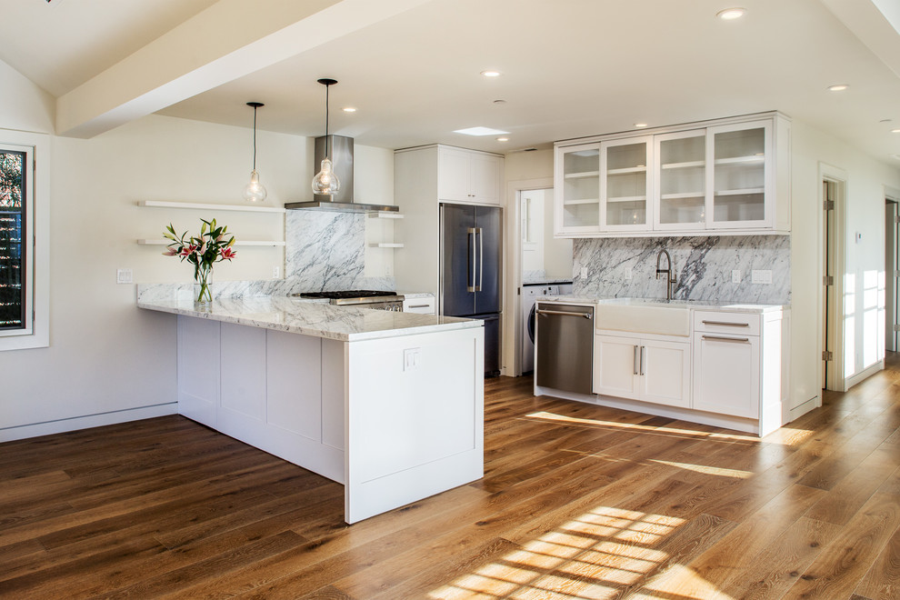 Photo of a mid-sized modern u-shaped open plan kitchen in San Francisco with a farmhouse sink, shaker cabinets, white cabinets, marble benchtops, white splashback, stone slab splashback, stainless steel appliances, medium hardwood floors, a peninsula and brown floor.