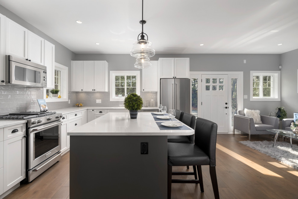 Design ideas for a mid-sized transitional l-shaped eat-in kitchen in Vancouver with a double-bowl sink, beaded inset cabinets, white cabinets, quartzite benchtops, grey splashback, ceramic splashback, stainless steel appliances, laminate floors, with island, brown floor and white benchtop.
