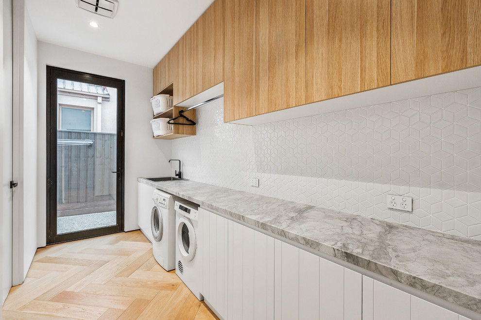 Photo of a large contemporary single-wall dedicated laundry room in Melbourne with a single-bowl sink, white cabinets, marble benchtops, white walls, light hardwood floors, a side-by-side washer and dryer and grey benchtop.