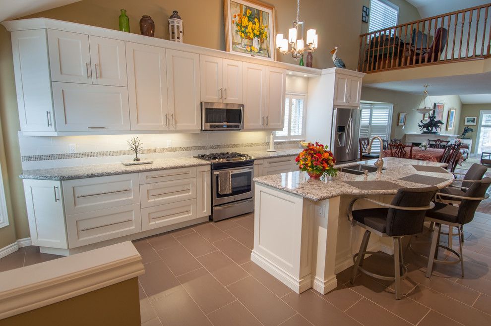Example of a mid-sized transitional galley ceramic tile eat-in kitchen design in Toronto with a double-bowl sink, shaker cabinets, white cabinets, granite countertops, beige backsplash, stone tile backsplash, stainless steel appliances and an island