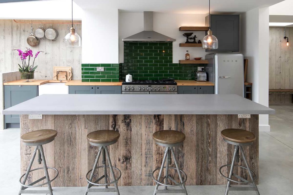 This is an example of a mid-sized industrial open plan kitchen in Sussex with a farmhouse sink, shaker cabinets, grey cabinets, wood benchtops, green splashback, subway tile splashback, stainless steel appliances, concrete floors, a peninsula, grey floor and grey benchtop.