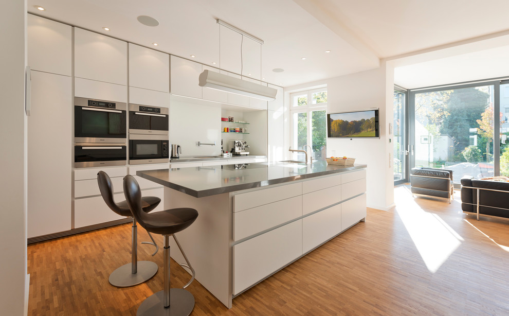Large contemporary open plan kitchen in Berlin with an undermount sink, flat-panel cabinets, white cabinets, white splashback, glass sheet splashback, stainless steel appliances, medium hardwood floors and with island.