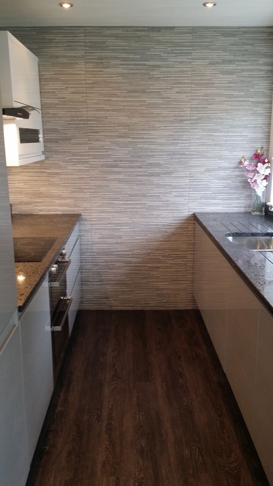 This is an example of a small contemporary galley separate kitchen in Cheshire with an integrated sink, flat-panel cabinets, white cabinets, granite benchtops, metallic splashback, glass sheet splashback, panelled appliances, vinyl floors and no island.