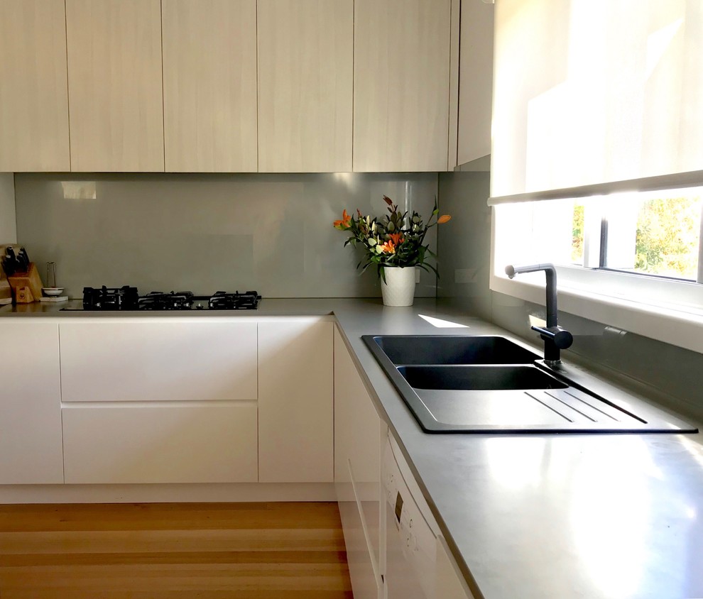 This is an example of a mid-sized contemporary l-shaped eat-in kitchen in Melbourne with a double-bowl sink, white cabinets, marble benchtops, metallic splashback, glass sheet splashback, black appliances, light hardwood floors, with island, brown floor and grey benchtop.